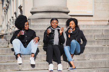 Fototapeta premium three beautiful and stylish dark-skinned girls with long hair standing in a city and drinking a coffee and use the phones