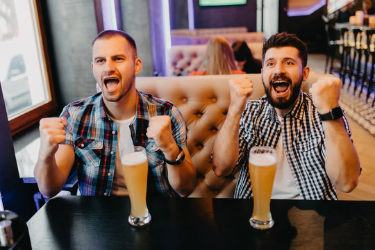 Fans two young men in plaid shirt at bar raised their hands with glasses of beer up and screaming, emotions from exciting game on TV