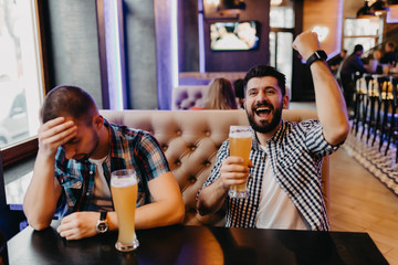 Two young friends fans of different team with lose and win emotions while drinking beer in mug at the pub watching football game
