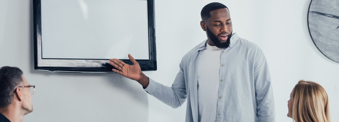 panoramic shot of smiling african american businessman pointing with hand at tv and talking with colleagues