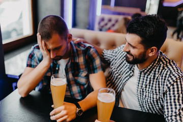 Consoling his depressed friend. Depressed young man sitting at the bar counter and holding head in hand while being consoled by his friend sitting near him