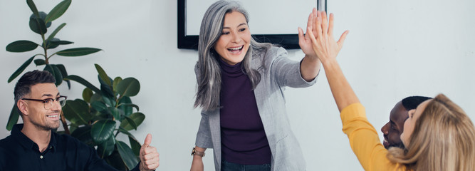 panoramic shot of smiling multicultural colleagues giving high five and showing like