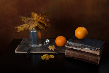 Two oranges, stack of vintage books, herbarium and pewter carafe with faded leaves on old black...