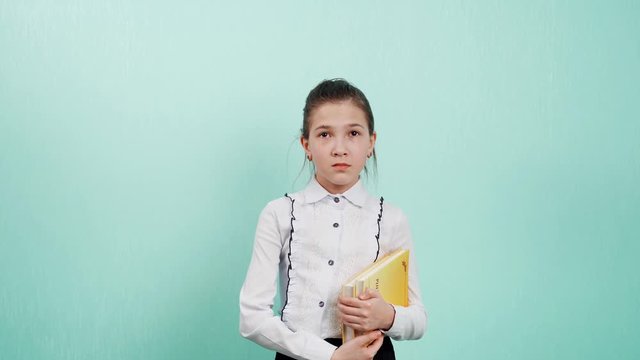 Portrait of a young girl with books in hand, putting hand in ear, asking someone to repeat or listen to bad news, isolated blue wall background. I don&rsquo;t hear you talking. All OK.