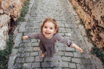 Pretty baby girl hold arms wide open and walk on a footpath on a medieval alley.
