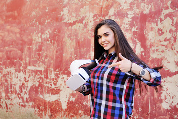 young cute girl with long hair in VR glasses in plaid shirt on red grunge wall background.