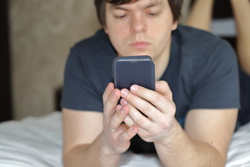 a dark- haired man is lying in bed in the morning with a phone and an e -book, dependence on gadgets in the morning, all the interests of a person in a smartphone