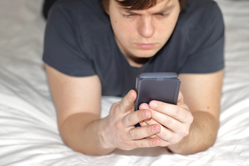 a dark- haired man is lying in bed in the morning with a phone and an e -book, dependence on gadgets in the morning, all the interests of a person in a smartphone