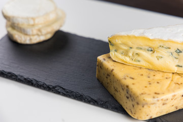 Fresh cheeses of different flavors with nuts on black chalkboard plate and wooden board on white...