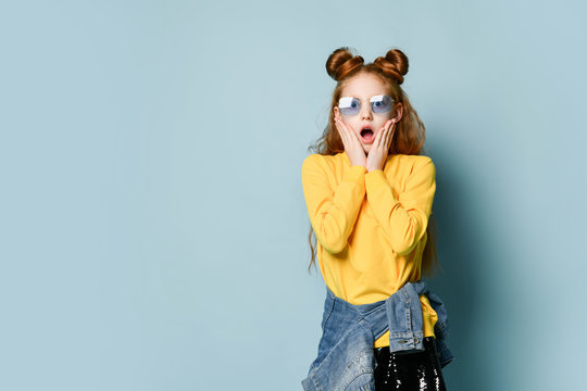 Amazed teen girl in blue smoked glasses and yellow sweatshirt is standing at free copy space covering cheeks with hands