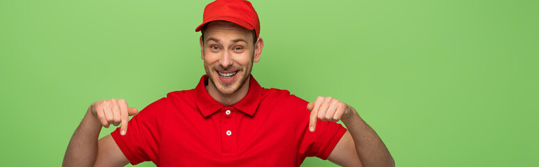 smiling delivery man in red uniform pointing with finger down isolated on green, panoramic shot