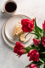 a slice of cake on a light plate, tulips, and black coffee on a light background