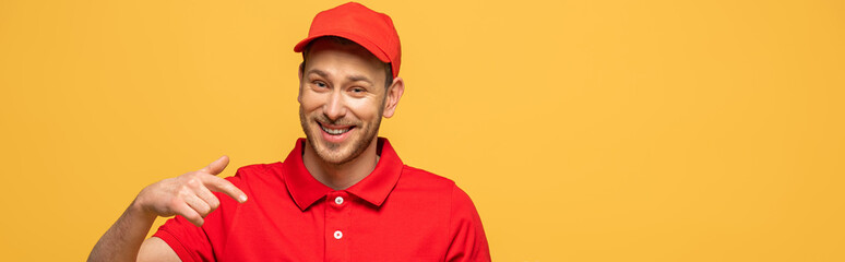 happy delivery man in red uniform pointing with finger isolated on yellow, panoramic shot