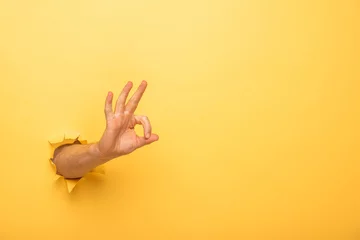 Fotobehang cropped view of man showing ok sign through yellow paper hole © LIGHTFIELD STUDIOS