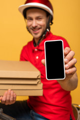 selective focus of happy delivery man in red uniform holding pizza boxes and smartphone with blank screen isolated on yellow