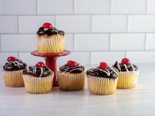 6 yellow cupcakes filled with custard with chocolate frosting and white drizzle with a cherry on top, decadent and delicious, sitting on a light sandstone counter with a white subway tile background.