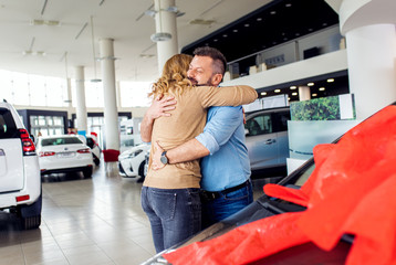 Smiling adult couple buying a new car from dealership, embracing each other. 