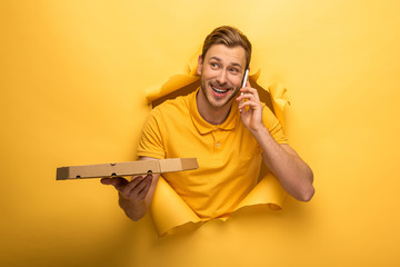 happy handsome man in yellow outfit talking on smartphone and holding pizza box in yellow paper hole