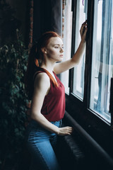 Close up portait of young sad, sensual, depressed, serious caucasian red headed gril with freckles standing near the window. Psychological condition concept