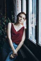 Close up portait of young sad, sensual, depressed, serious caucasian red headed gril with freckles standing near the window. Psychological condition concept