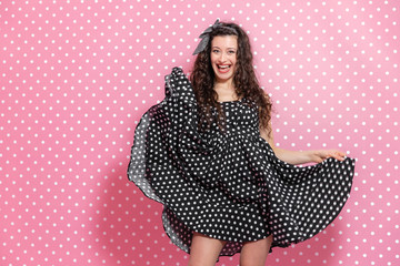 Pinup curly girl is making windy dress, posing, grinning at the camera.