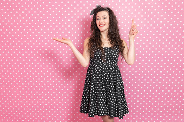 Beautiful young girl with kinky hair wearing nice black polka-dots dress is pointing at copyspace...