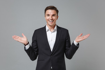 Excited smiling young business man in classic black suit shirt posing isolated on grey wall background studio portrait. Achievement career wealth business concept. Mock up copy space. Spreading hands.