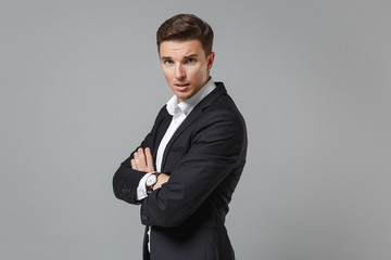 Obraz na płótnie Canvas Side view of shocked young business man in classic black suit shirt isolated on grey background. Achievement career wealth business concept. Mock up copy space. Holding hands crossed, looking camera.