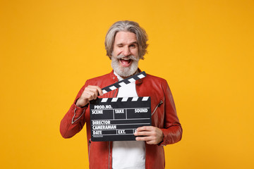 Blinking elderly gray-haired mustache bearded man in red leather jacket isolated on yellow orange background. People lifestyle concept. Mock up copy space. Hold classic black film making clapperboard.