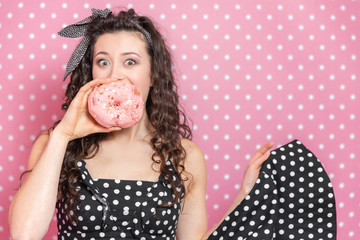 Enchanting girl is holding a doughnut in front of her mouth, raising the eyebrows and keeping eyes widely opened.