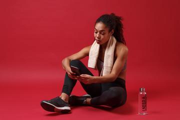 Smiling young african american woman in sportswear working out isolated on red background. Sport exercises healthy lifestyle concept. Sit with towel over her neck, water bottle, using mobile phone.