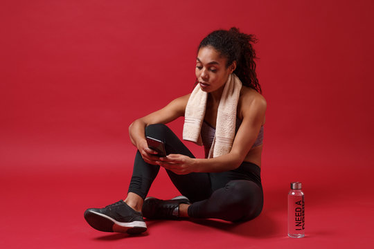 Smiling young african american woman in sportswear working out isolated on red background. Sport exercises healthy lifestyle concept. Sit with towel over her neck, water bottle, using mobile phone.
