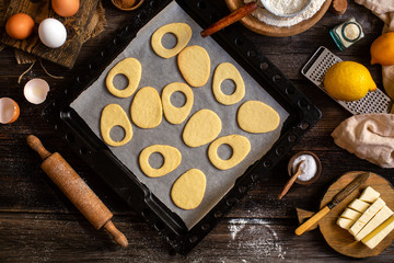 overhead shot of baked egg shaped easter cookies on baking tray on rustic wooden table with flour, eggs, butter, lemon. process of baking cookies. easter card