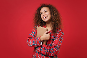 Pensive young african american girl in pajamas homewear posing while resting at home isolated on red background in studio. Relax good mood lifestyle concept. Mock up copy space. Hold book, looking up.