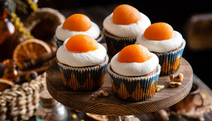 easter carrot cupcakes with white cream and slice of apricot on top (egg style decorated cupcakes) on wooden cake stand on rustic table with willow, oranges, nuts, spices