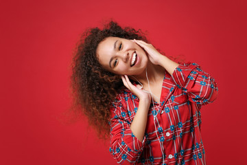 Cheerful young african american girl in pajamas homewear posing while resting at home isolated on red background. Relax good mood lifestyle concept. Mock up copy space. Listen music with earphones.