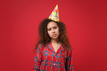Displeased young african american girl in pajamas homewear, birthday hat posing while resting at home isolated on red background. Relax good mood lifestyle concept. Mock up copy space. Looking camera.