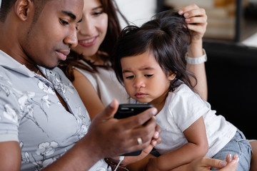 Caucasian woman and african-american man and little daughter watching together photos from the rest on a smartphone sitting on cozy armchair. Concept of strong family relationships and summer memories