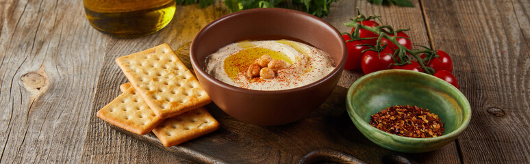 Crackers, bowls with hummus and spices on cutting board, cherry tomatoes, jar of olive oil on wooden background, panoramic shot
