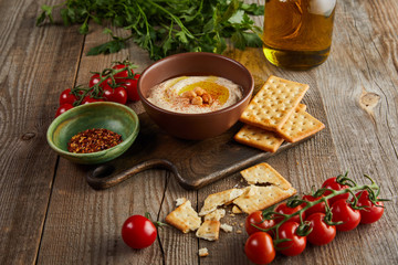 Crackers, bowls with hummus and spices on cutting board, vegetables and jar of olive oil on wooden background