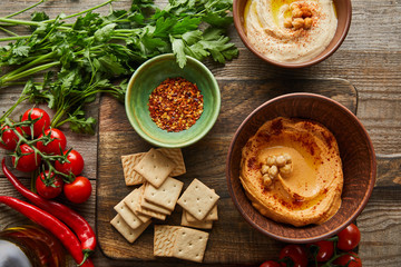 Top view of crackers, bowls with spices and hummus on cutting board with vegetables and parsley on wooden background