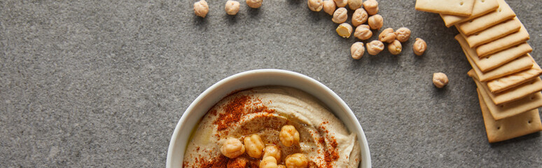 Top view of bowl with delicious hummus, chickpea and crackers on grey background, panoramic shot