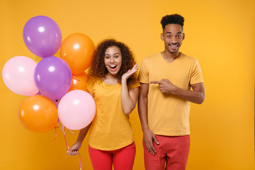 Excited friends couple african american guy girl in casual clothes isolated on yellow background. Birthday holiday party concept. Celebrate hold colorful air balloons spreading hands, pointing aside.