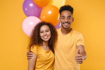Smiling young friends couple african american guy girl in casual clothes isolated on yellow background. Birthday holiday party concept. Celebrate hold colorful air balloons hugging showing thumb up.