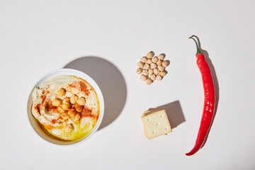 Top view of bowl with hummus near crackers, chili pepper and chickpea on grey