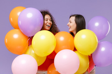 Fototapeta na wymiar Laughing two european african american women friends in knitted sweaters isolated on violet purple background. Birthday holiday party, people emotions concept. Celebrating hold colorful air balloons.
