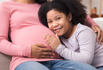 Adorable black girl hugging her pregnant mom's tummy, smiling at camera