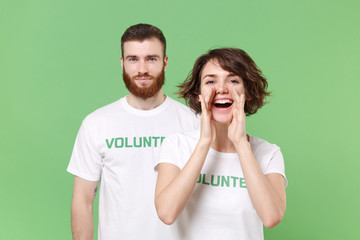 Funny two friends couple in white volunteer t-shirt isolated on pastel green background. Voluntary free work assistance help charity grace teamwork concept. Screaming with hands gesture near mouth.