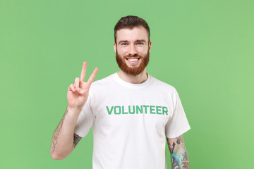 Smiling young bearded man in white volunteer t-shirt isolated on pastel green wall background...