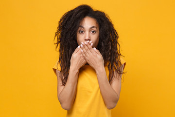 Shocked young african american woman girl in casual t-shirt posing isolated on yellow orange wall background studio portrait. People lifestyle concept. Mock up copy space. Covering mouth with hands.